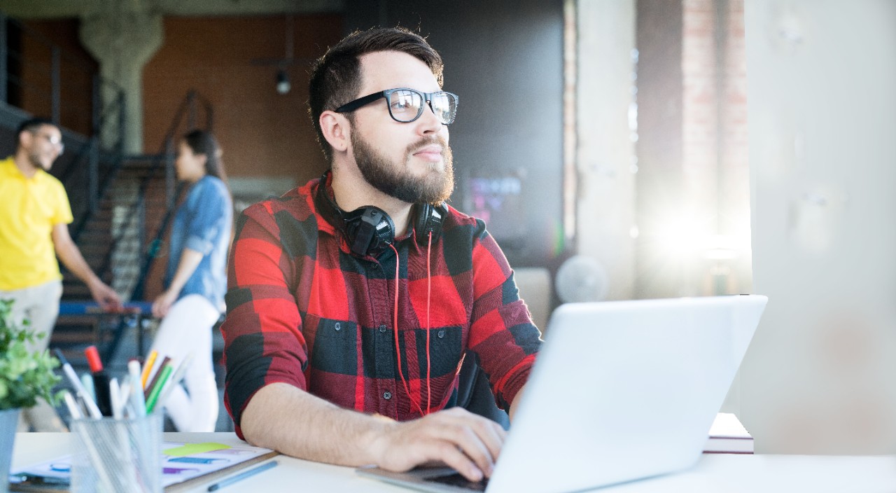 Portrait of modern bearded man wearing glasses and red shirt using laptop looking away pensively in sunlit office of IT developers team, copy space